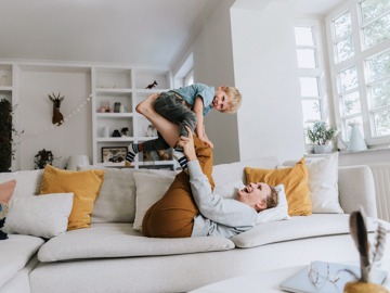 Eine Mutter liegt auf der Couch und stemmt spielerisch ihren jungen Sohn mit den Füßen in die Höhe. Beide lachen